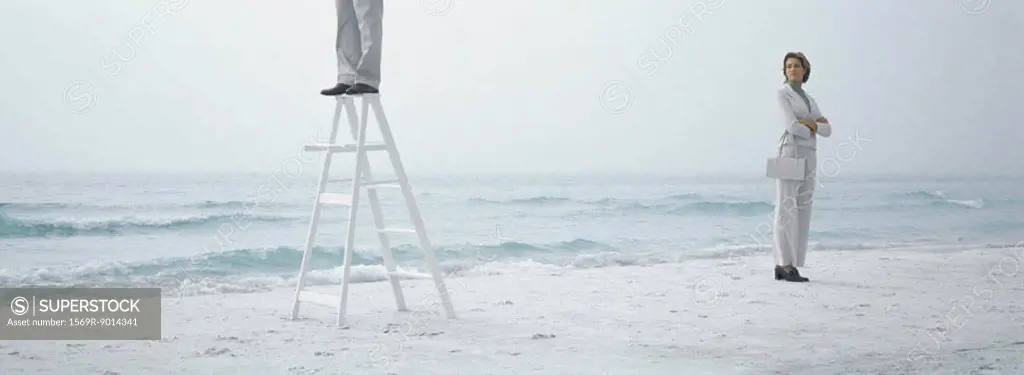 Woman standing on beach with arms crossed, looking toward man standing on ladder