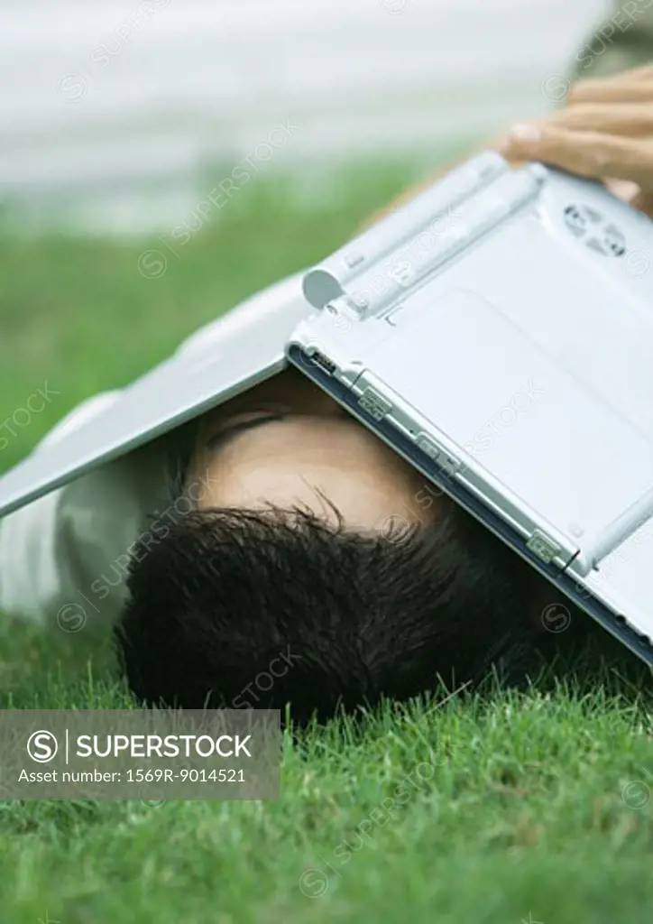 Man lying in grass with laptop covering face