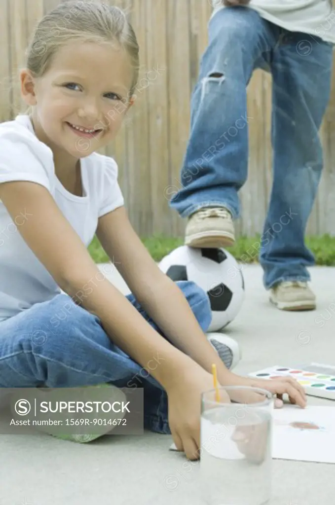 Girl sitting on driveway, painting, boy with foot on soccerball in background