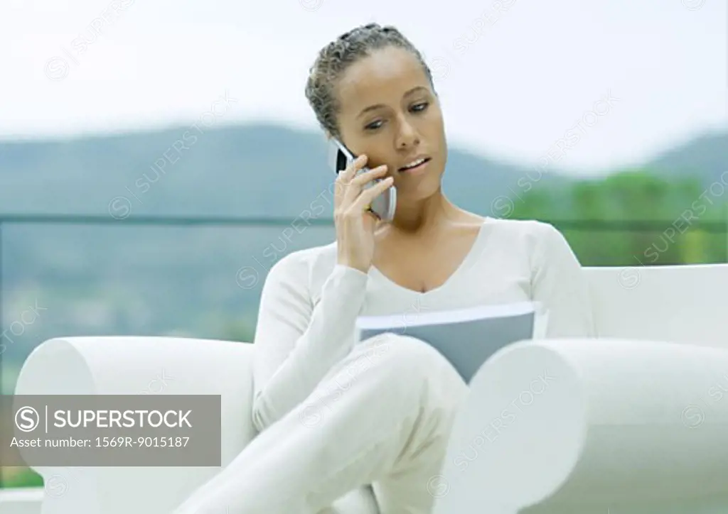 Woman sitting outdoors in armchair, using cell phone and looking at document