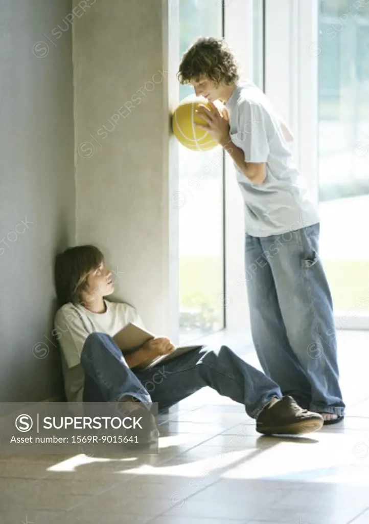 Boy sitting on floor studying, friend holding basketball, talking to him