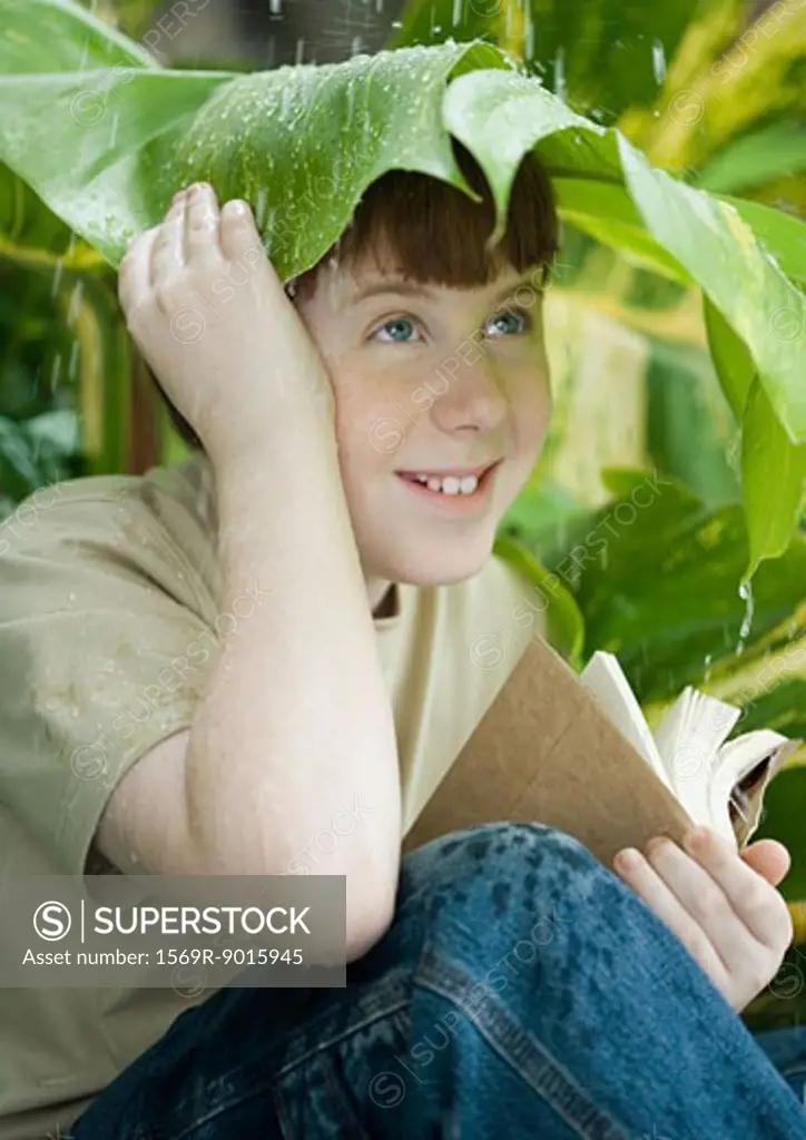 Boy with book sitting under leaf in rain