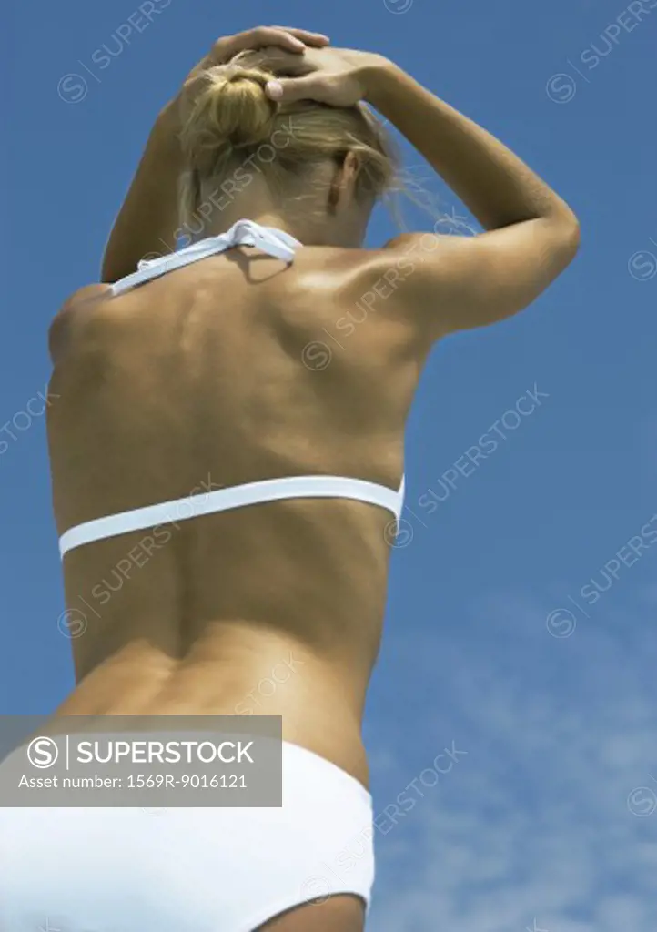 Woman standing in sun in bikini, hands behind head, low angle rear view