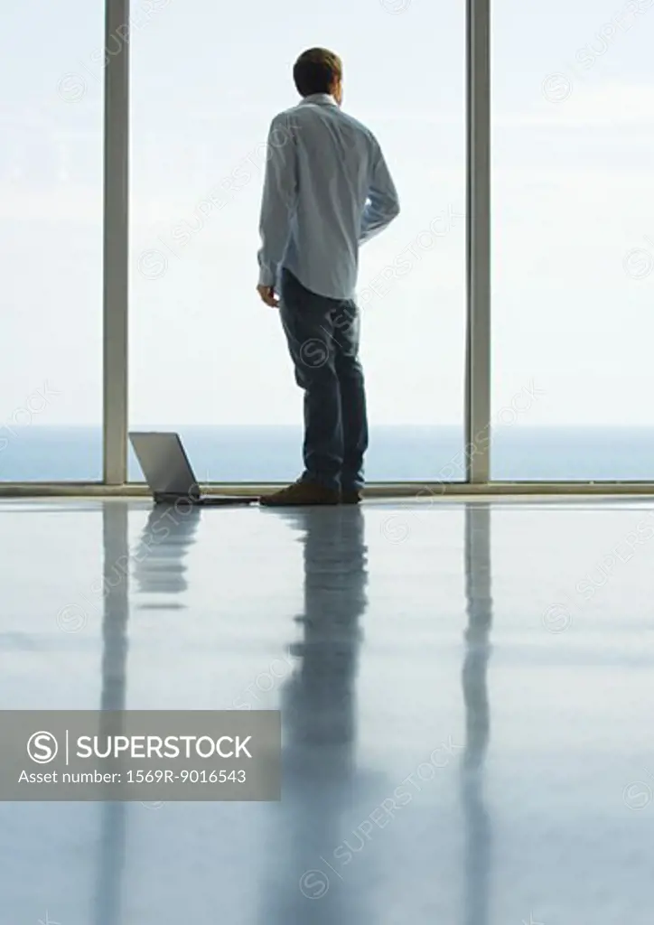 Young man standing by window, laptop on floor, full length
