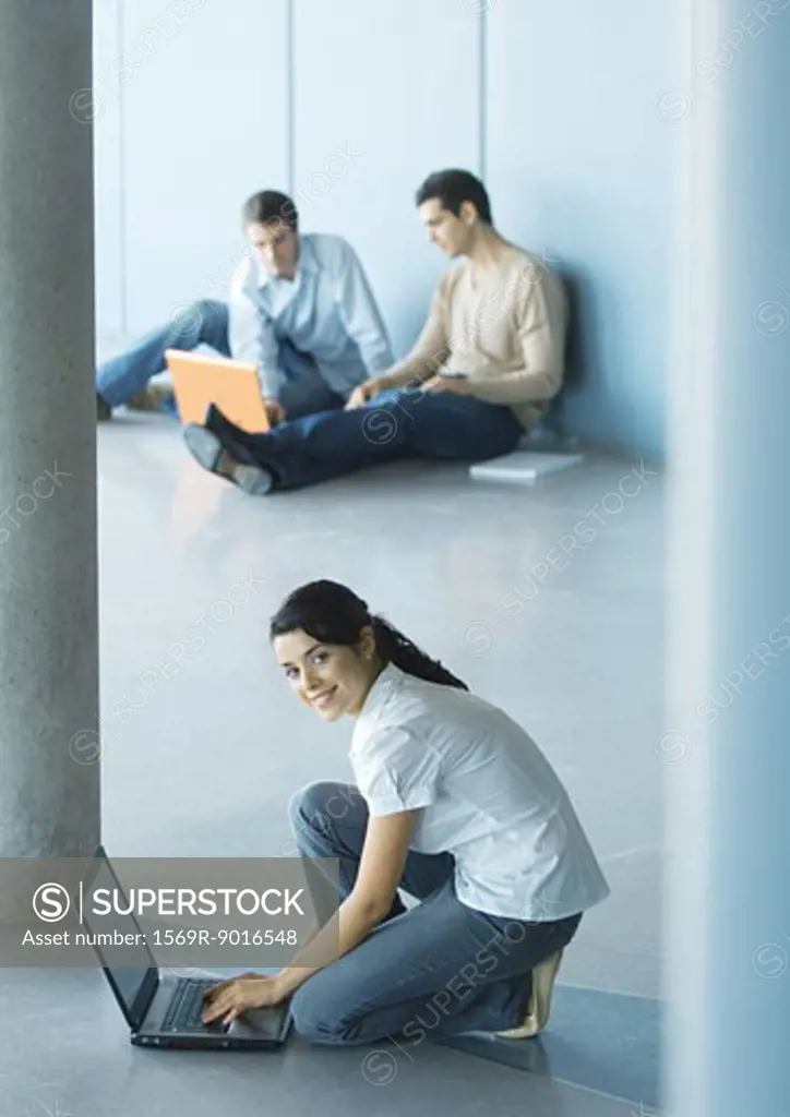 Young woman crouching sitting on floor, using laptop, men sitting in background