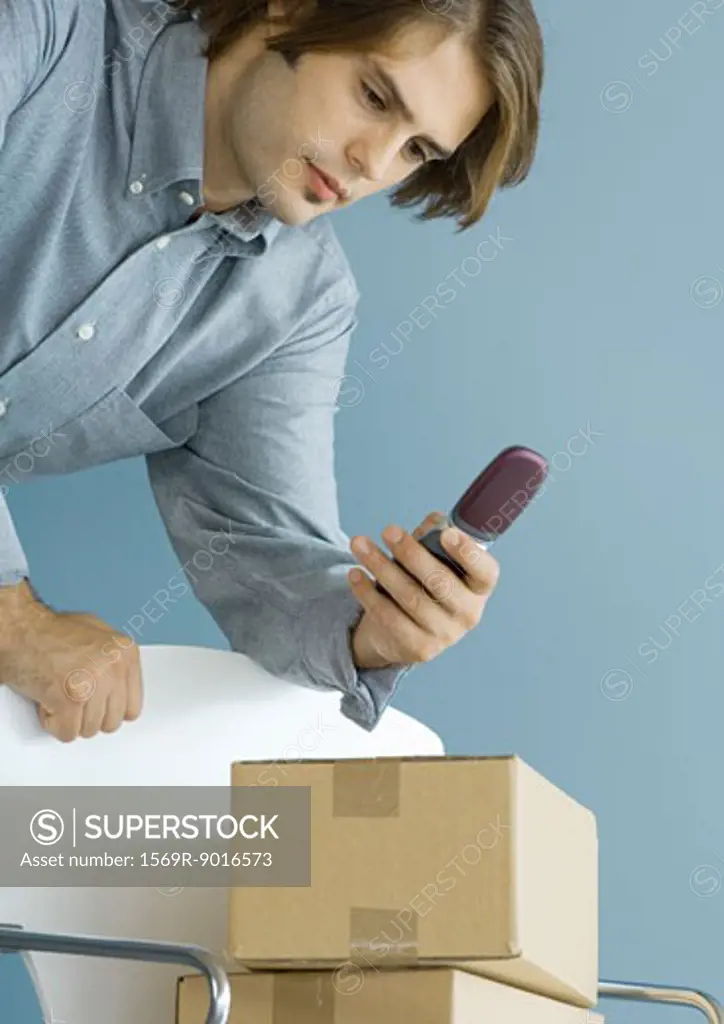 Man leaning against chair, looking at cell phone, cardboard boxes stacked on chair