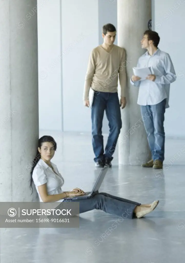 Young woman sitting on floor, using laptop, two men standing behind her