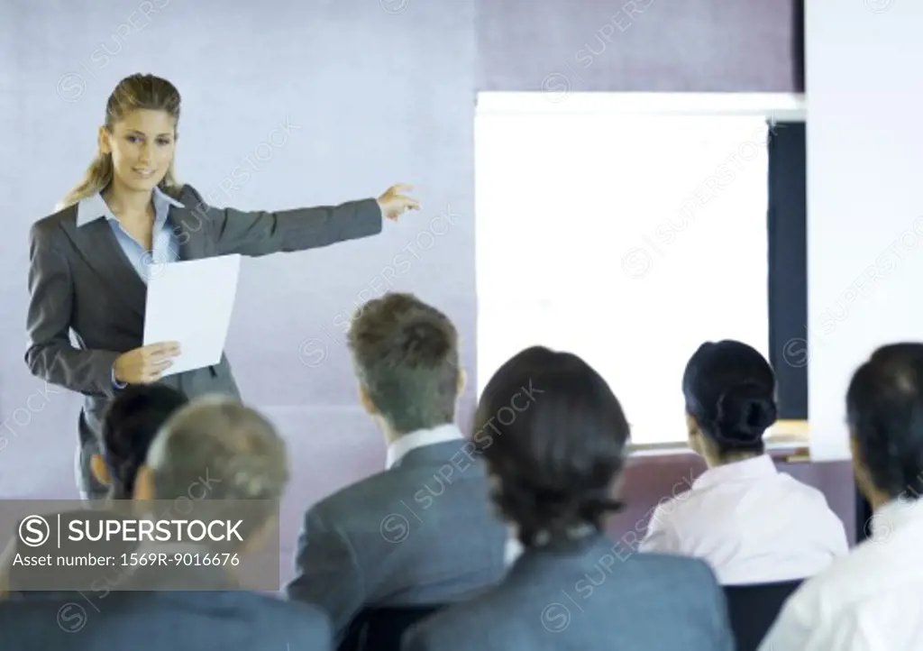 Executives sitting in seminar, woman standing facing group, gesturing to screen