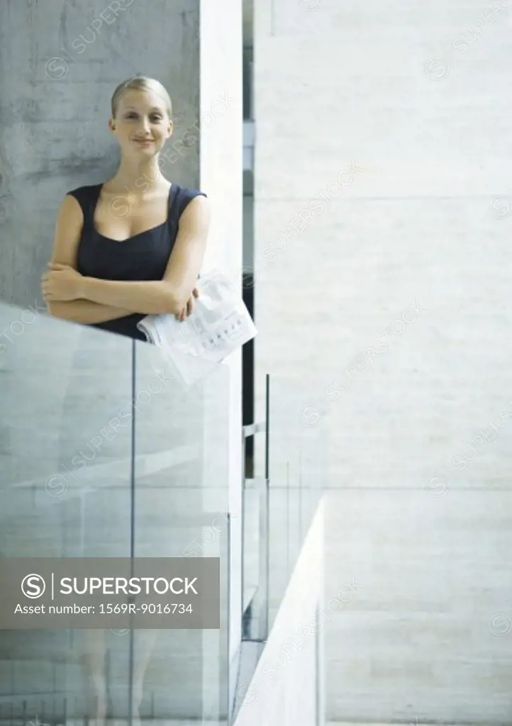 Businesswoman standing next to glass guard rail, holding newspaper, portrait
