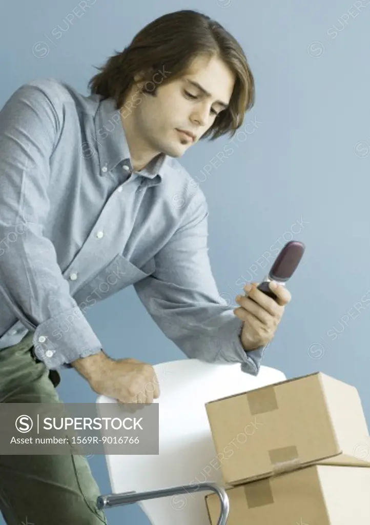 Man looking at cell phone, leaning on chair piled with cardboard boxes