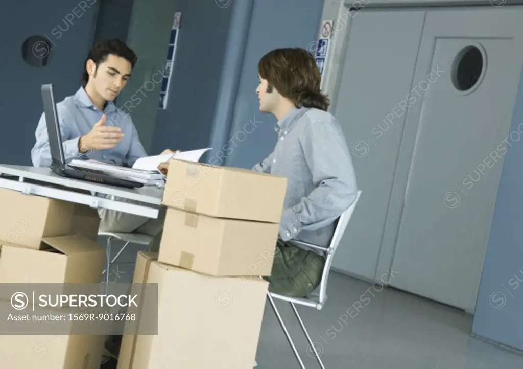 Two male colleagues talking across desk, cardboard boxes in foreground