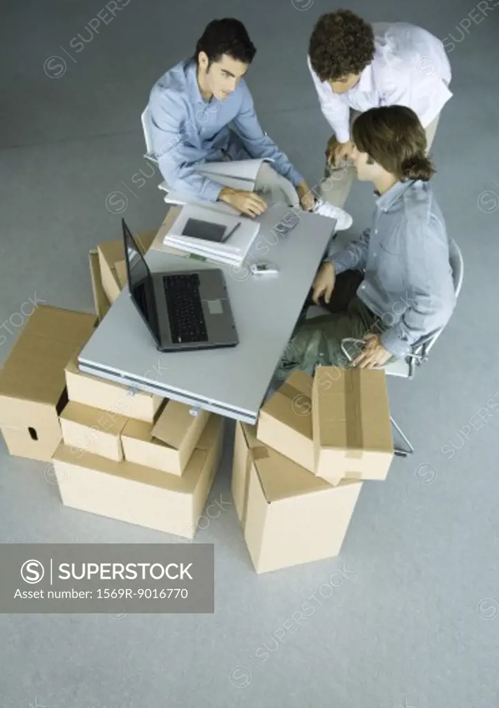 Three male colleagues grouped around cell phone on desk, surrounded by cardboard boxes