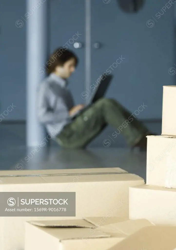 Young man sitting on floor using laptop, cardboard boxes in foreground