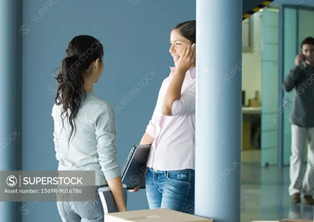 Two young women standing together near cardboard box, one using cell phone and holding laptop