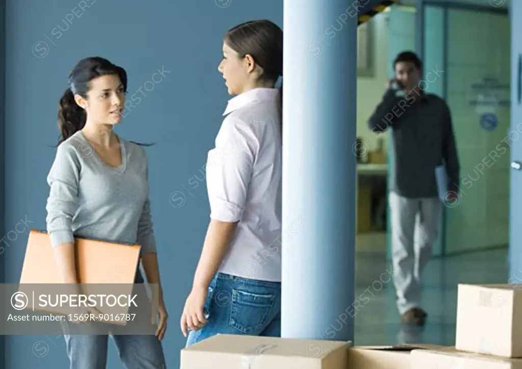 Two young women standing face to face talking, man in background using cell phone