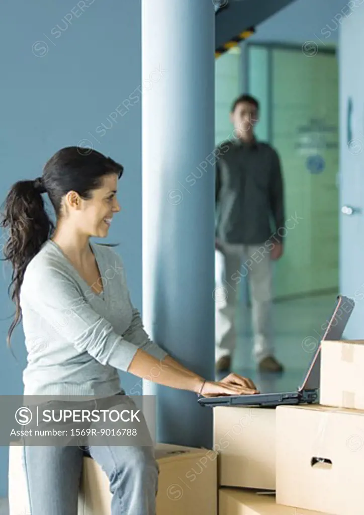 Woman sitting on cardboard box, using laptop, man standing in background