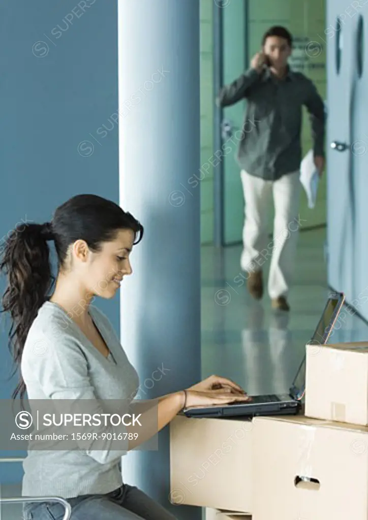 Woman sitting, using laptop on cardboard boxes, man approaching in background