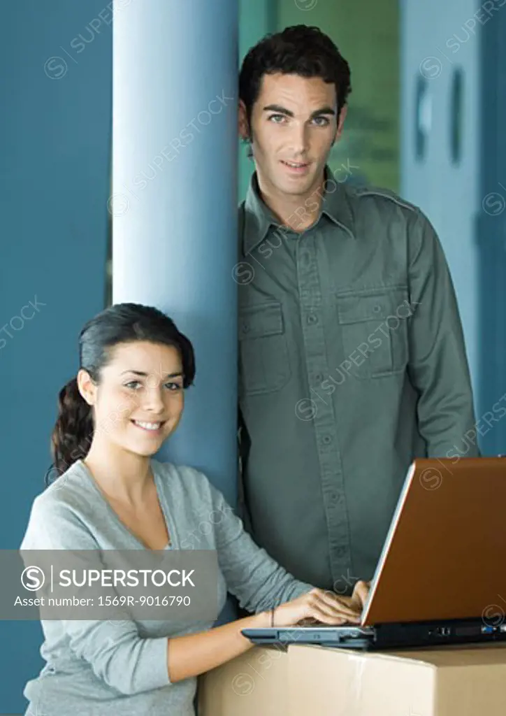 Woman sitting, using laptop on cardboard box, man standing next to her