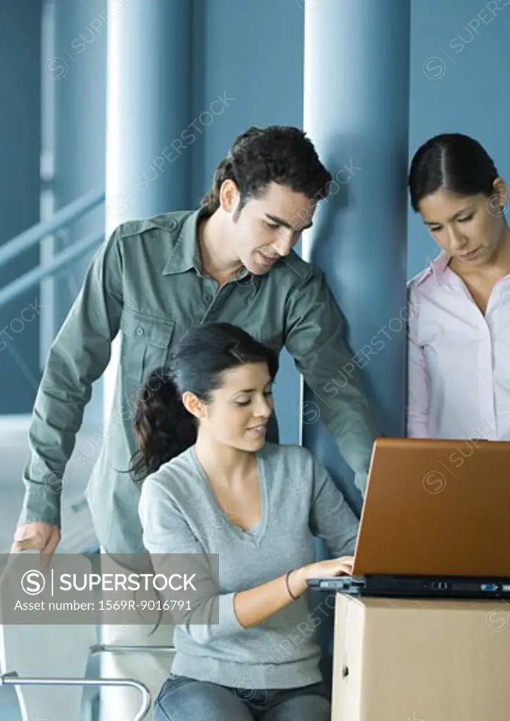 Woman sitting, using laptop on cardboard box, two colleagues looking over her shoulder