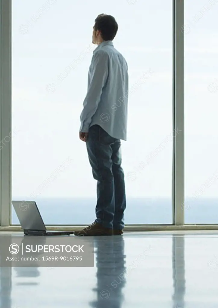 Man standing, looking out bay window, laptop on floor next to him
