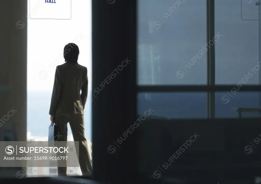 Woman standing with briefcase, looking out bay window, in silhouette