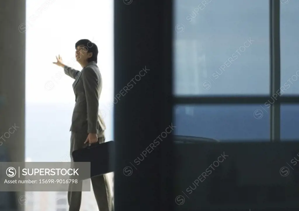 Woman standing with briefcase, next to bay window with arm out, looking over shoulder