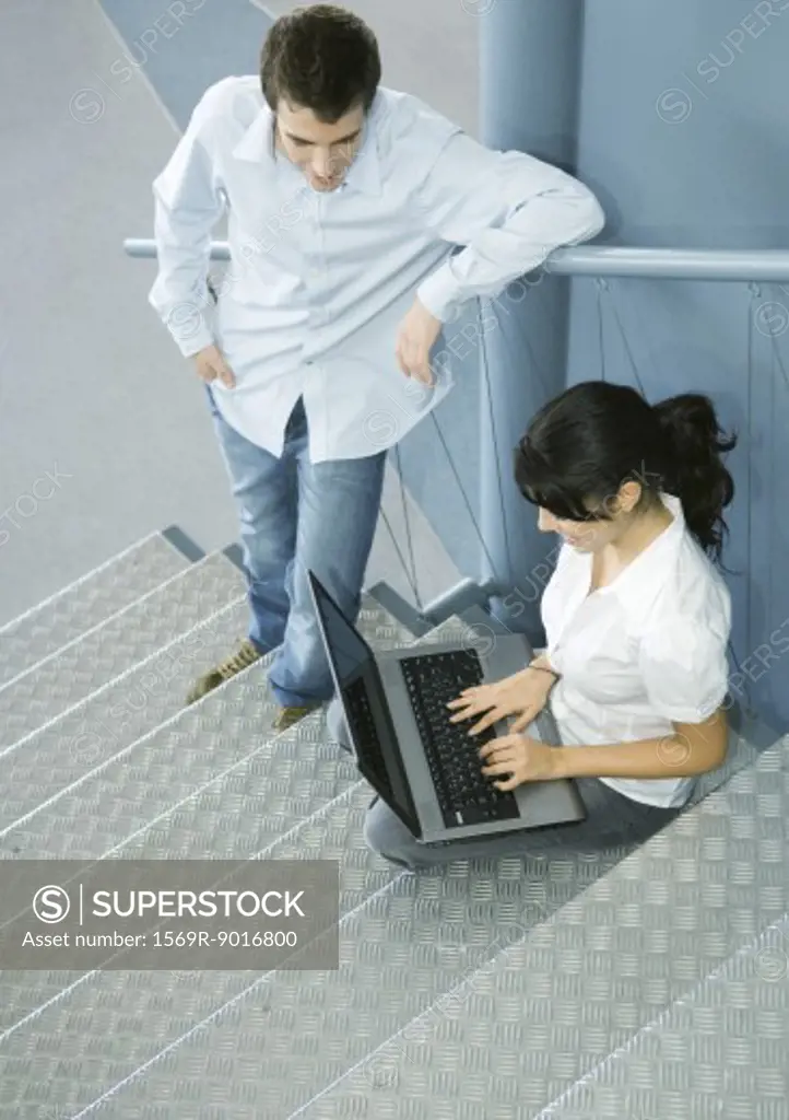 Two colleagues talking, one sitting on stairs with laptop