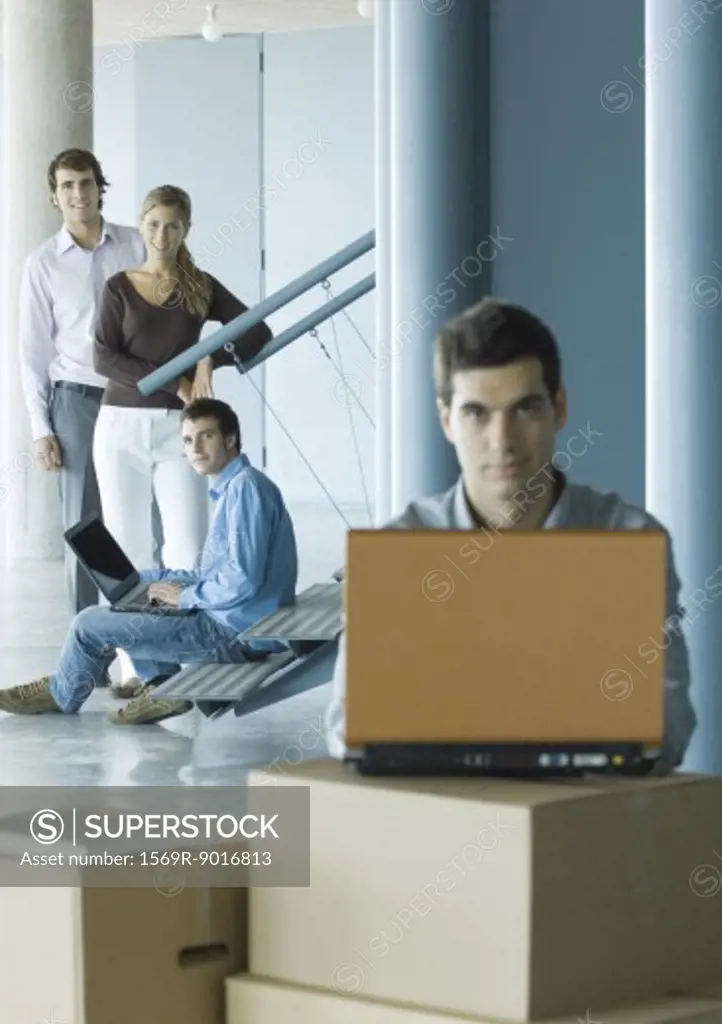 Man using laptop on stack of cardboard boxes, focus on colleagues standing in background