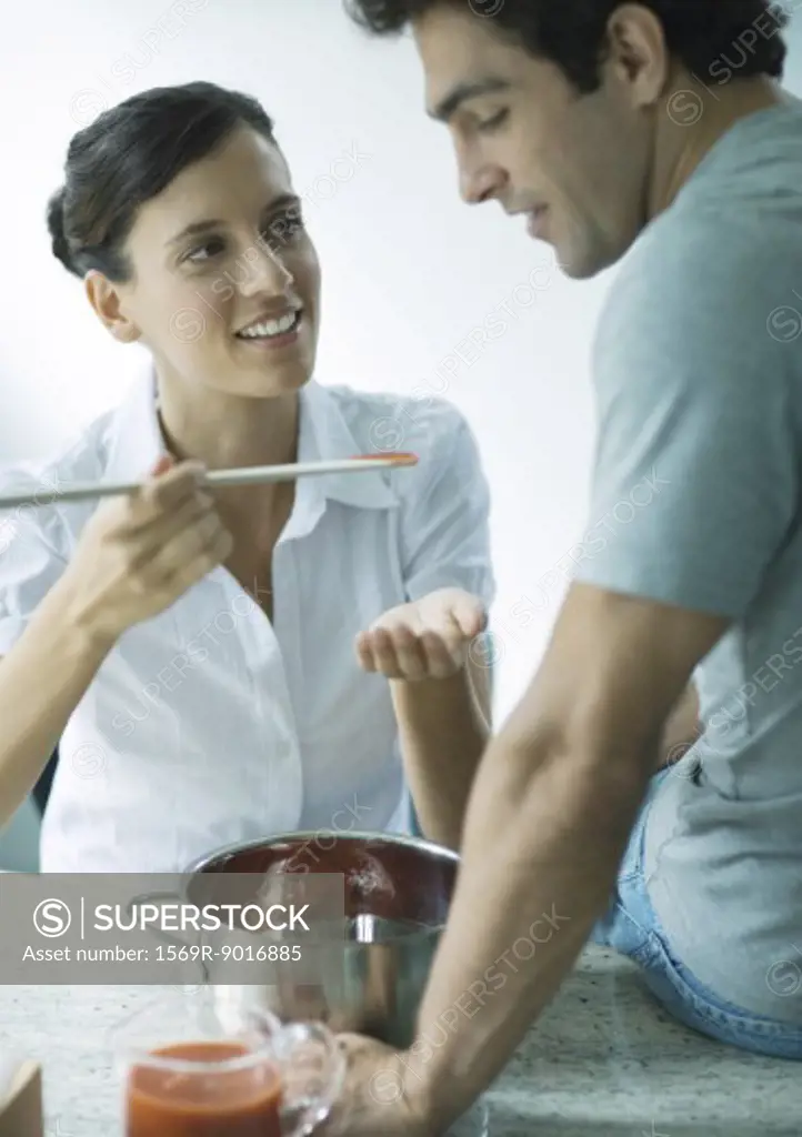 Woman holding up spoon of tomato sauce for man to taste