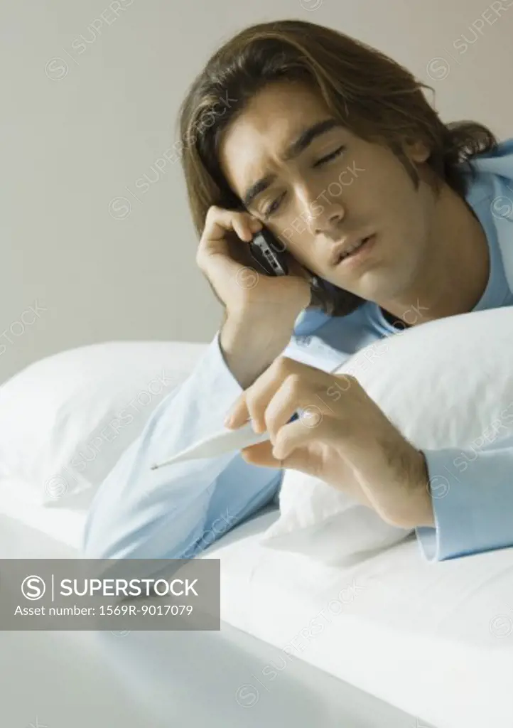 Man lying in bed, using cell phone and holding up thermometer