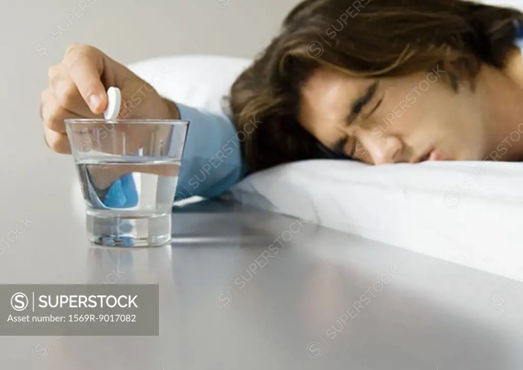 Man lying in bed, squinting eyes shut and holding effervescent medicine over glass of water