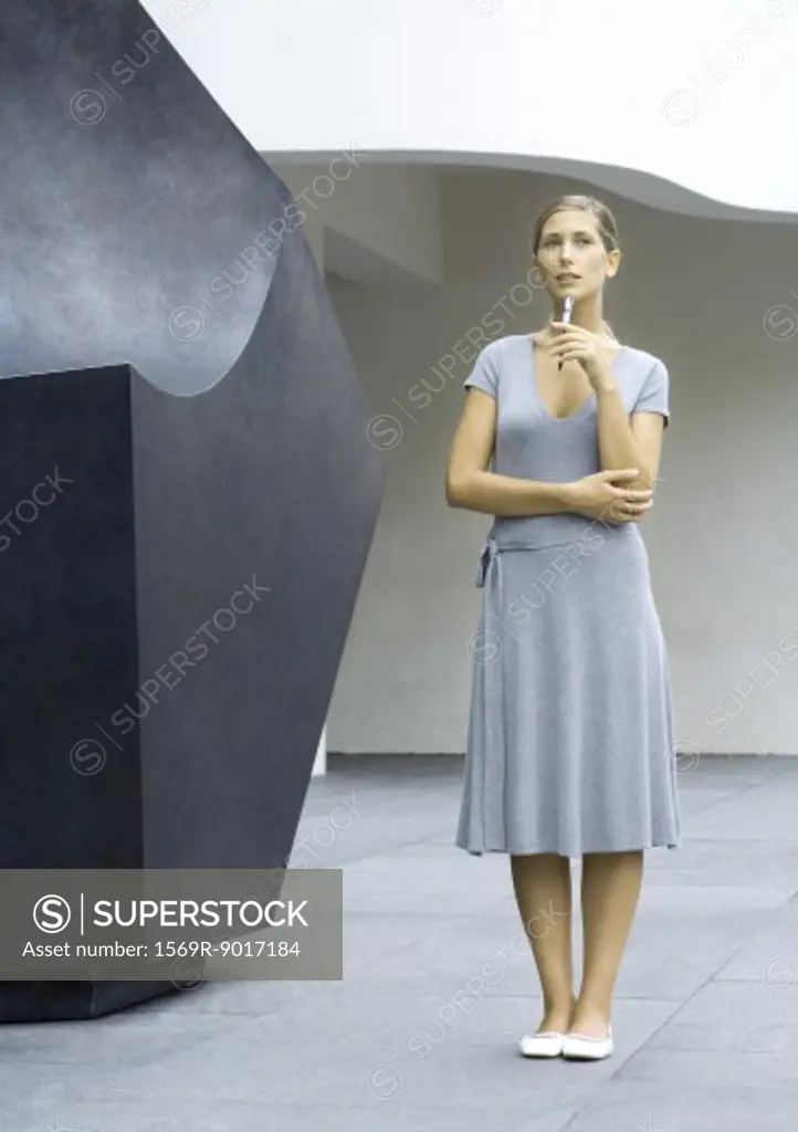 Woman standing next to sculpture, holding pen to chin, full length