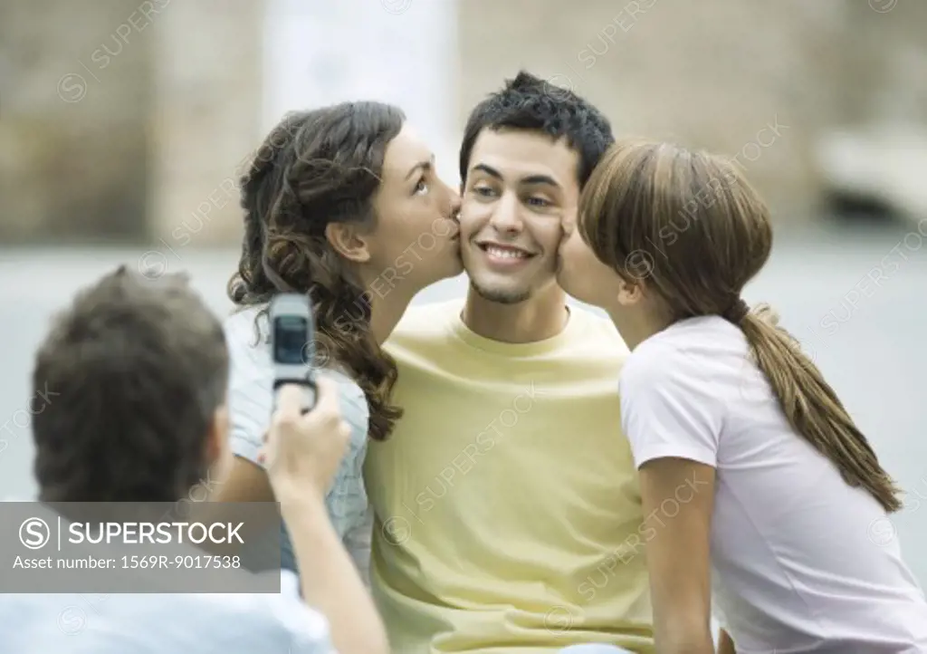 Teen girls kissing boy's cheeks while second boy takes photo