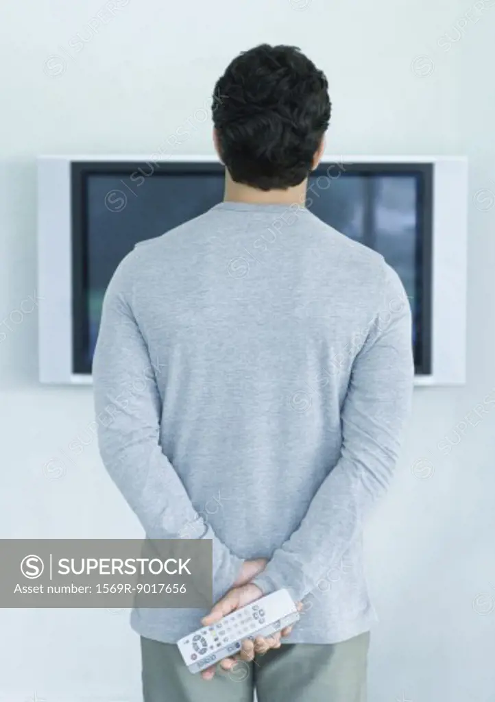 Man standing in front of widescreen TV, holding two remote controls, rear view