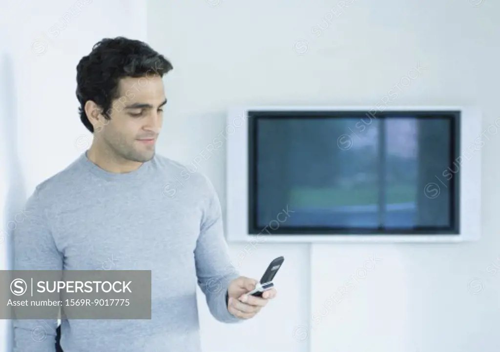 Man standing, looking at cell phone, widescreen TV on wall in background