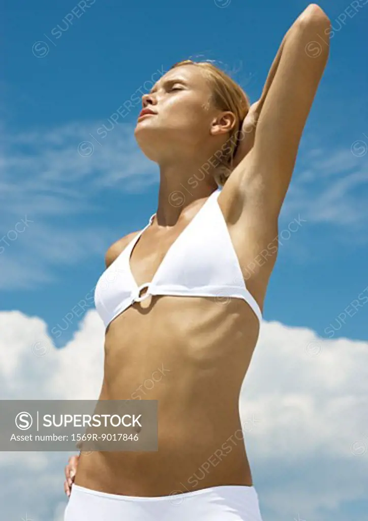 Woman wearing bikini, stretching with hand behind head, sky in background