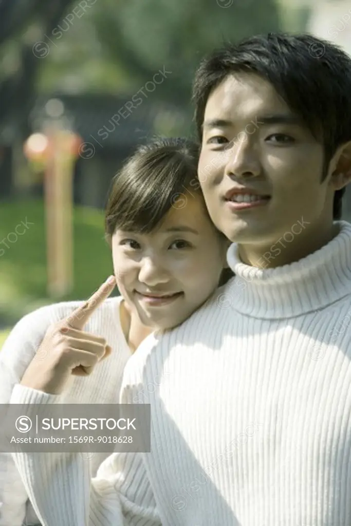 Young couple, woman resting head on man's shoulder, man pointing to woman, both smiling at camera