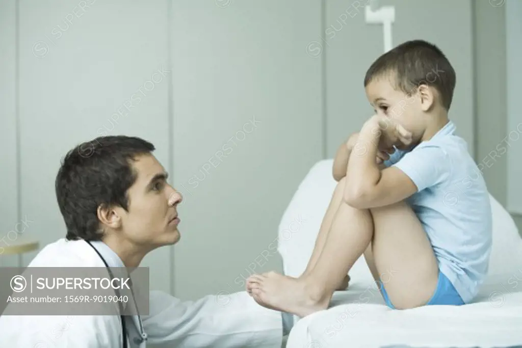 Boy sitting on examination table with knees up, doctor looking up at boy, profile