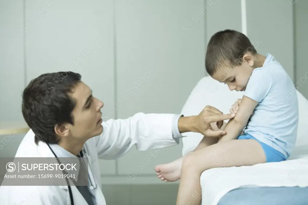 Boy sitting on examination table, doctor pointing to boy's arm