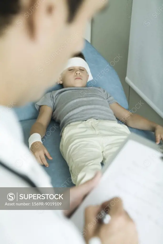 Boy lying on examination table with bandage on forehead and arm, doctor writing on clipboard