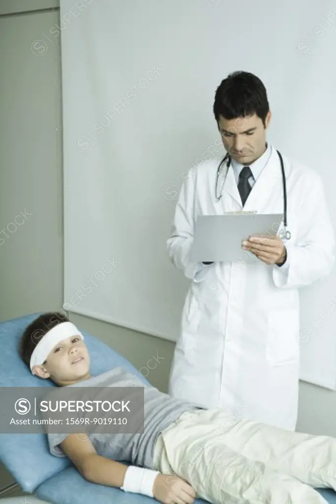 Boy lying on examination table with bandage on forehead and arm, doctor writing on clipboard