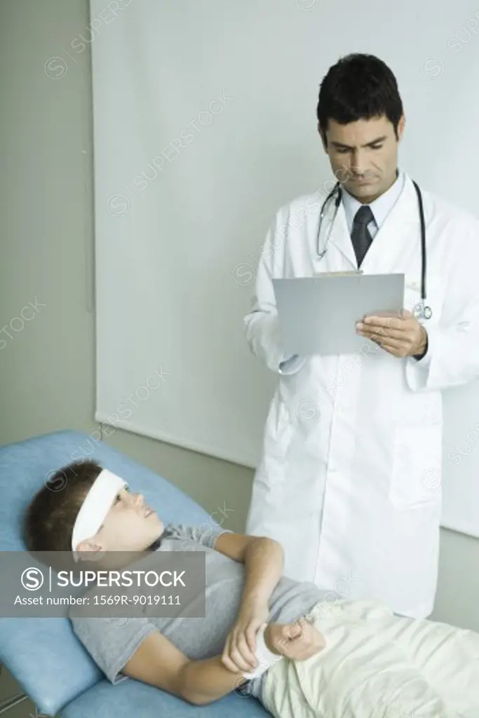 Boy lying on examination table with bandage on forehead, holding arm, doctor writing on clipboard