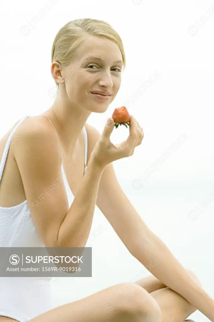 Teenage girl wearing bathing suit, holding up strawberry, smiling