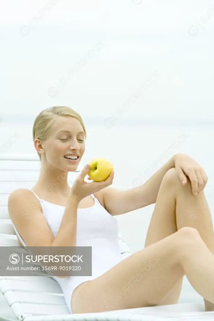 Teenage girl wearing bathing suit, sitting in lounge chair, eating apple