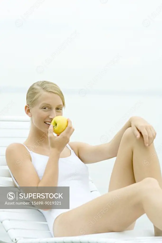 Teenage girl wearing bathing suit, sitting in lounge chair, eating apple