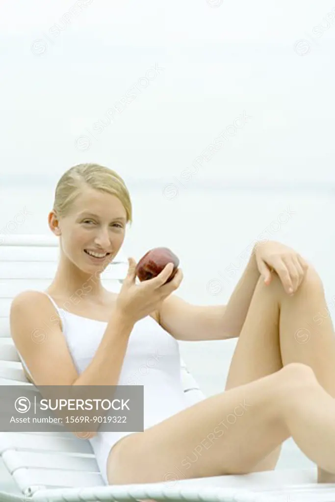 Teenage girl wearing bathing suit, sitting in lounge chair, holding apple