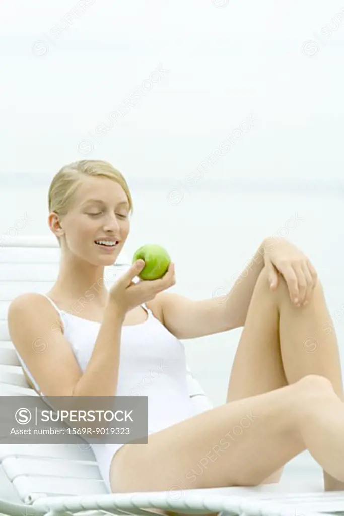 Teenage girl wearing bathing suit, sitting in lounge chair, holding apple