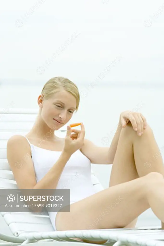 Teenage girl wearing bathing suit, sitting in lounge chair, holding carrot stick