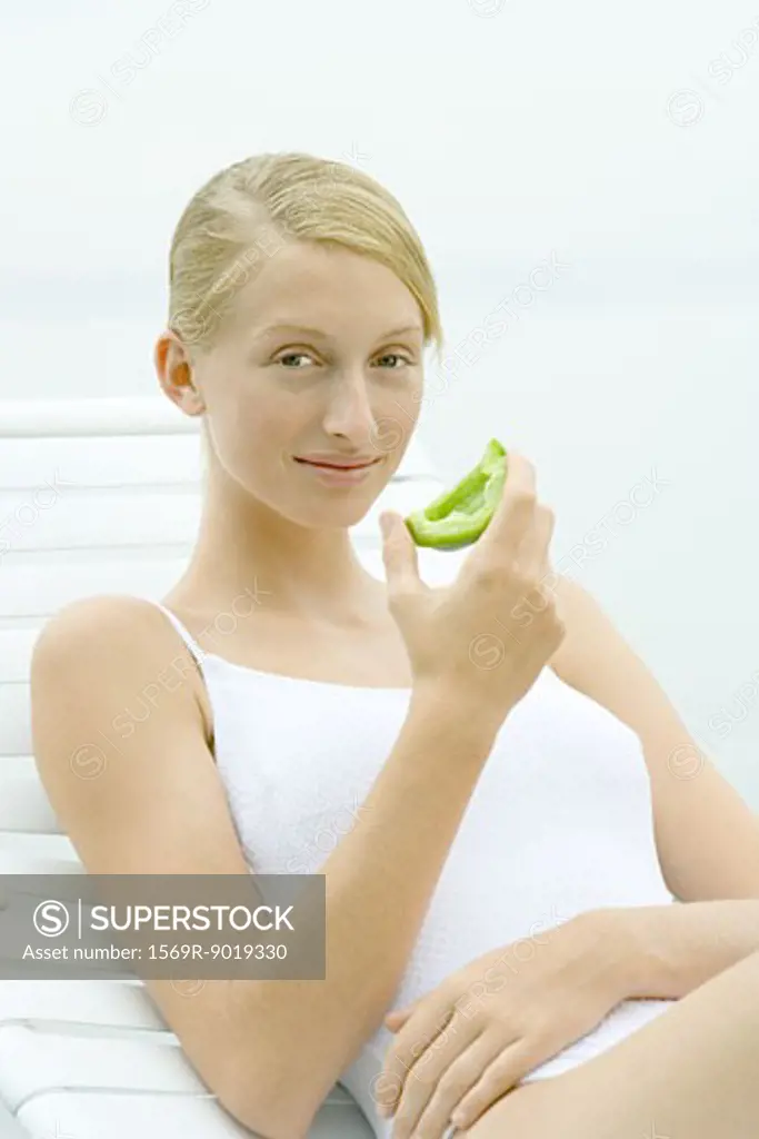 Teenage girl wearing bathing suit, sitting in lounge chair, holding piece of green pepper