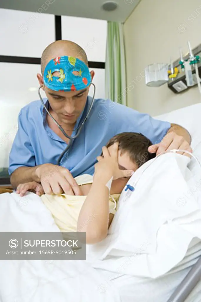 Boy lying in hospital bed holding head, doctor listening to boy with stethoscope